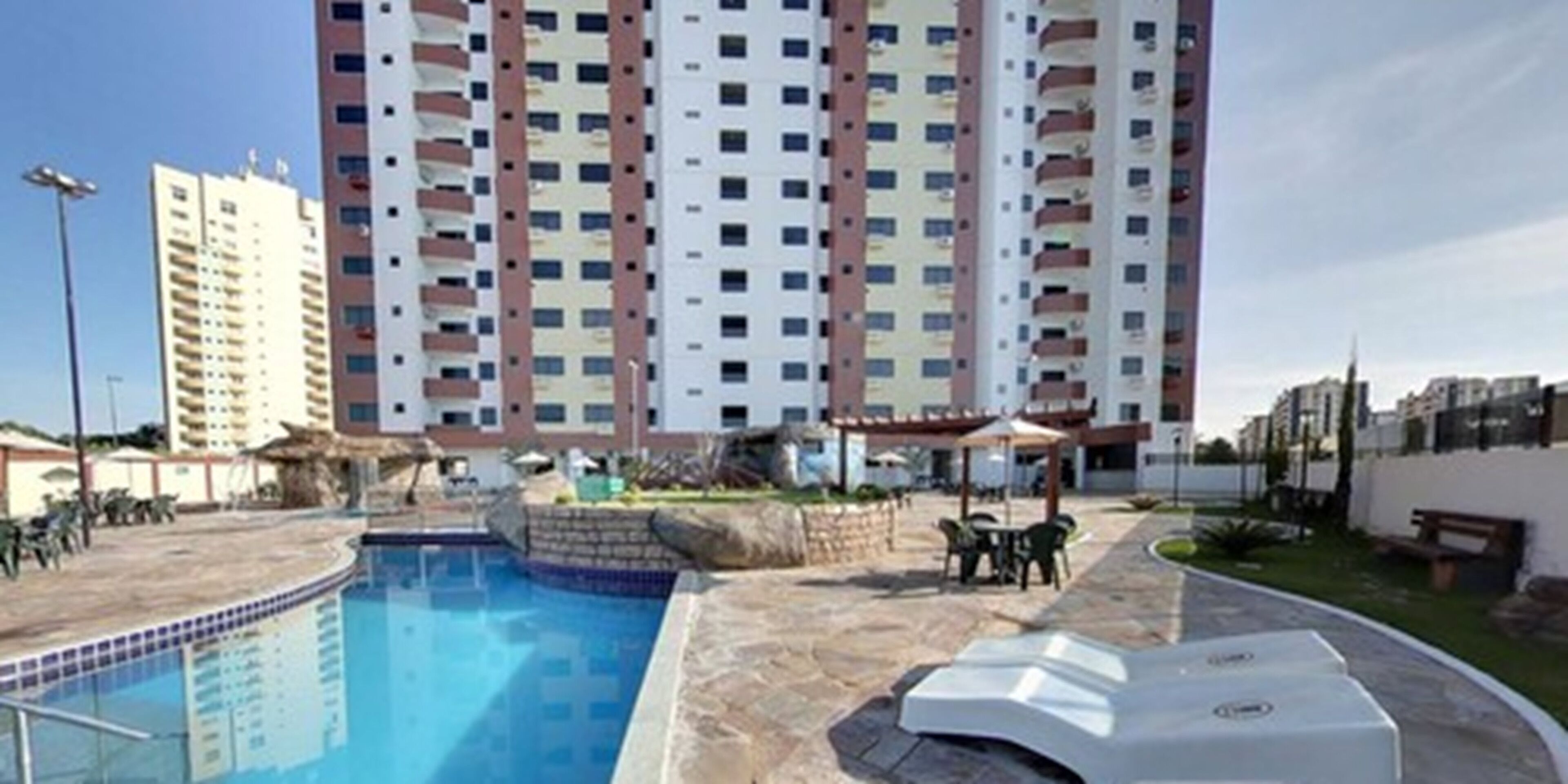 Ein Schwimmbecken mit einer Steindeck, umgeben von einer Terrasse mit Gartenmöbeln. Im Hintergrund ein mehrstöckiges Wohngebäude mit Balkonen.