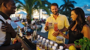 Uma área de bar com um bartender, um cliente segurando uma bebida e outro cliente. Há garrafas e copos no balcão.