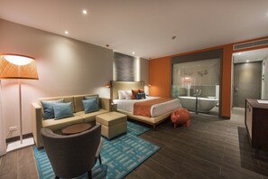 A modern hotel room with a sofa, a bed, a round coffee table, and a bathroom with a bathtub.
