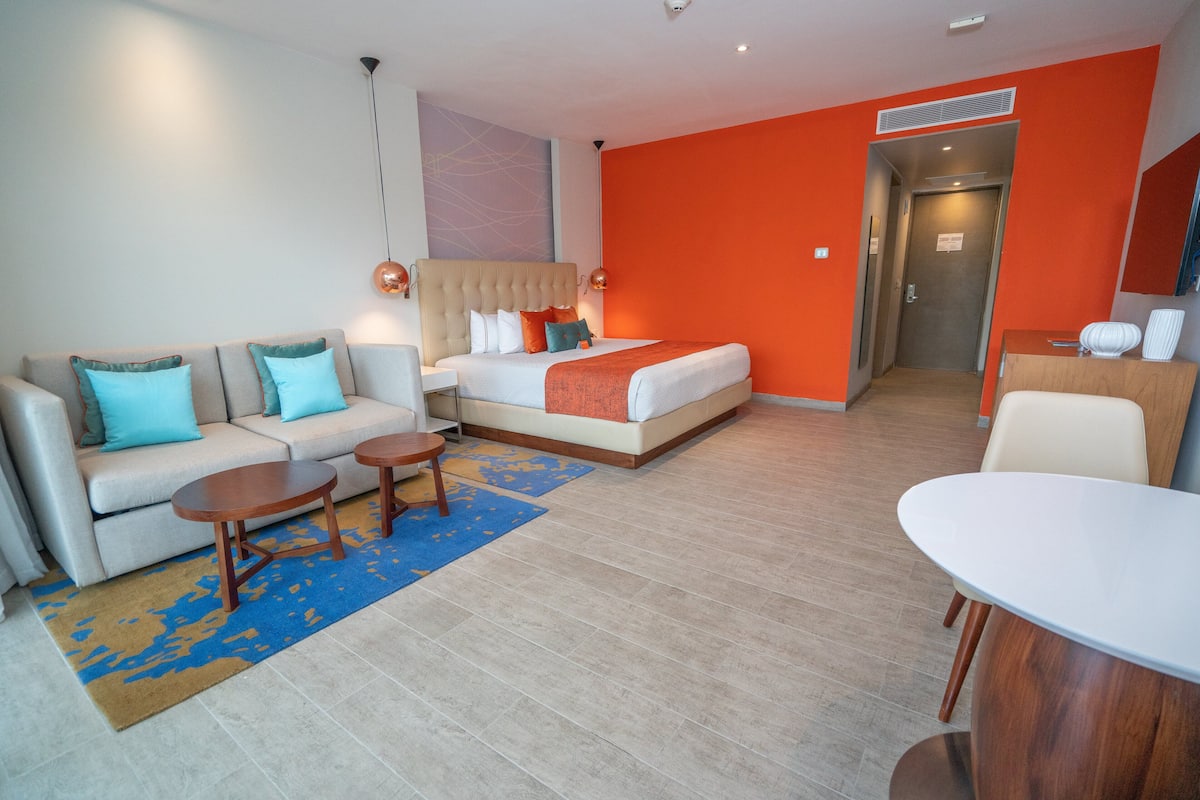 Ein modernes Hotelzimmer mit einer Couch, einem Bett mit orangefarbener Bettwäsche, einem Esstisch und einer kleinen Küchenzeile.