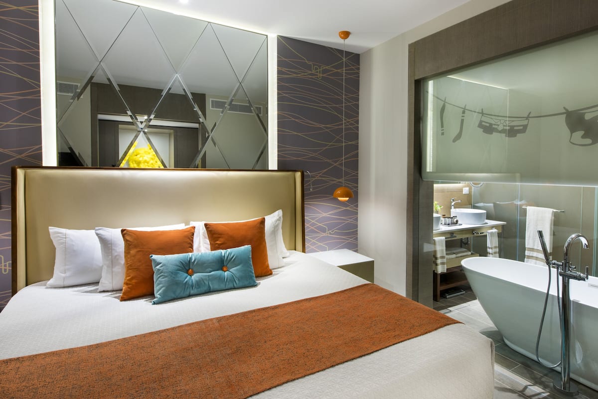 Ein modernes Hotelzimmer mit einem großen Bett, einem Badezimmer mit einer Badewanne und einem Spiegel, der den Raum wiederspiegelt.