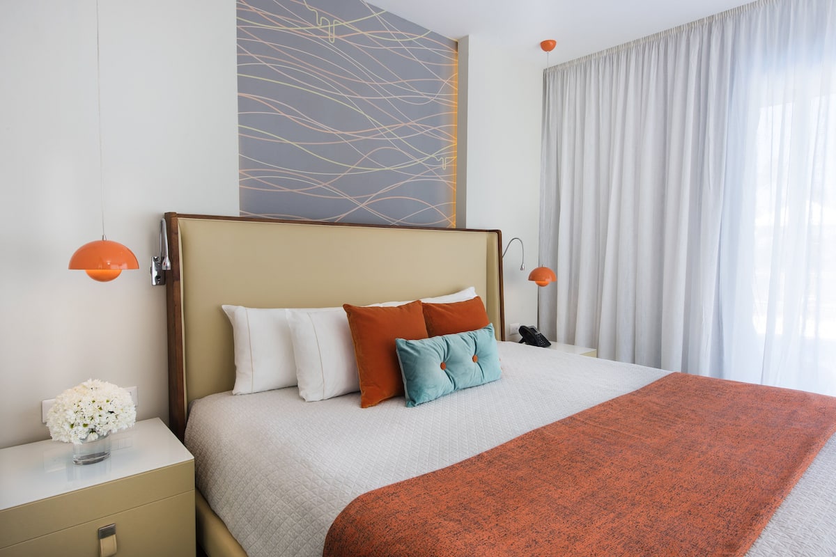 Ein ordentlich bezogenes Bett mit einem beige farbenen Kopfteil, weißen und orangefarbenen Kissen und einer roten Decke. Ein Nachttisch mit Blumen in einer Vase und einer Lampe steht daneben.