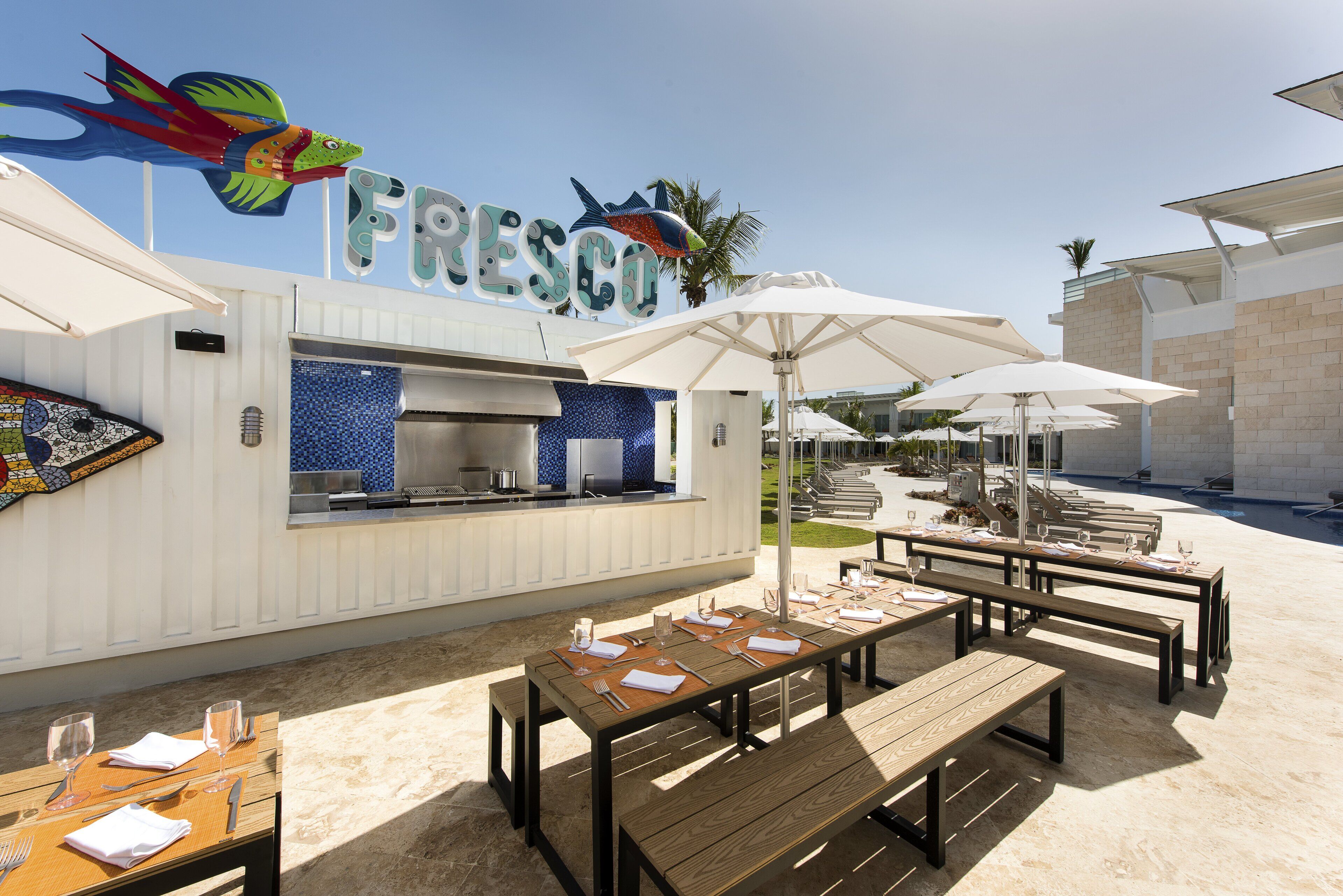 Un espace extérieur aménagé pour les repas, avec des tables et des parasols, un coin cuisine et un panneau décoré d’un motif coloré représentant un poisson.