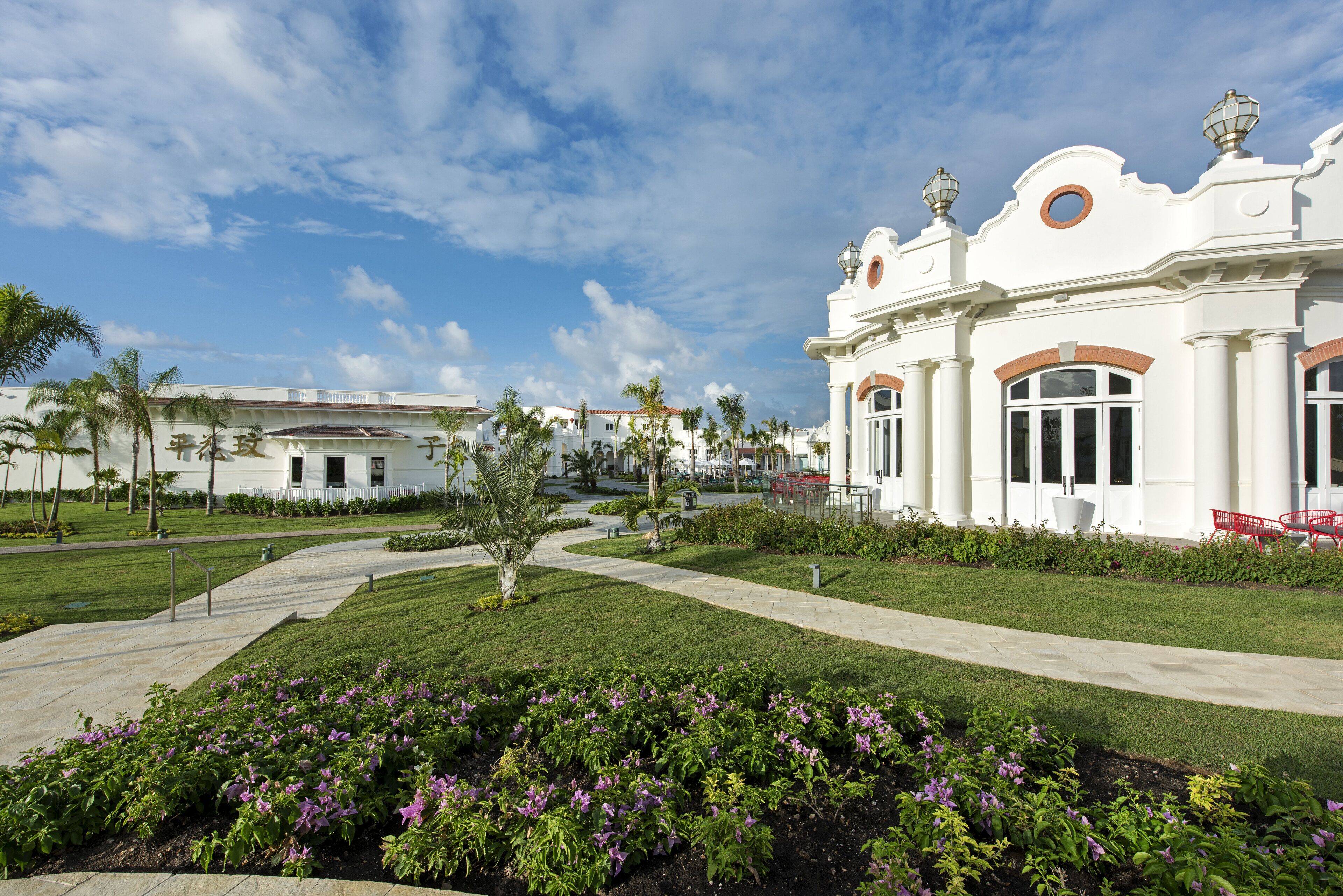A white building with arched windows and a circular window on the second floor. There is a pathway leading to the building, surrounded by green grass and purple flowers.