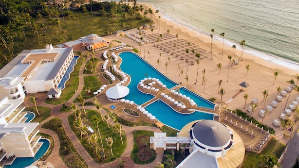 Vista aérea de um resort com uma grande piscina central, várias piscinas menores, uma área de praia com espreguiçadeiras e uma linha costeira bordeada por palmeiras.