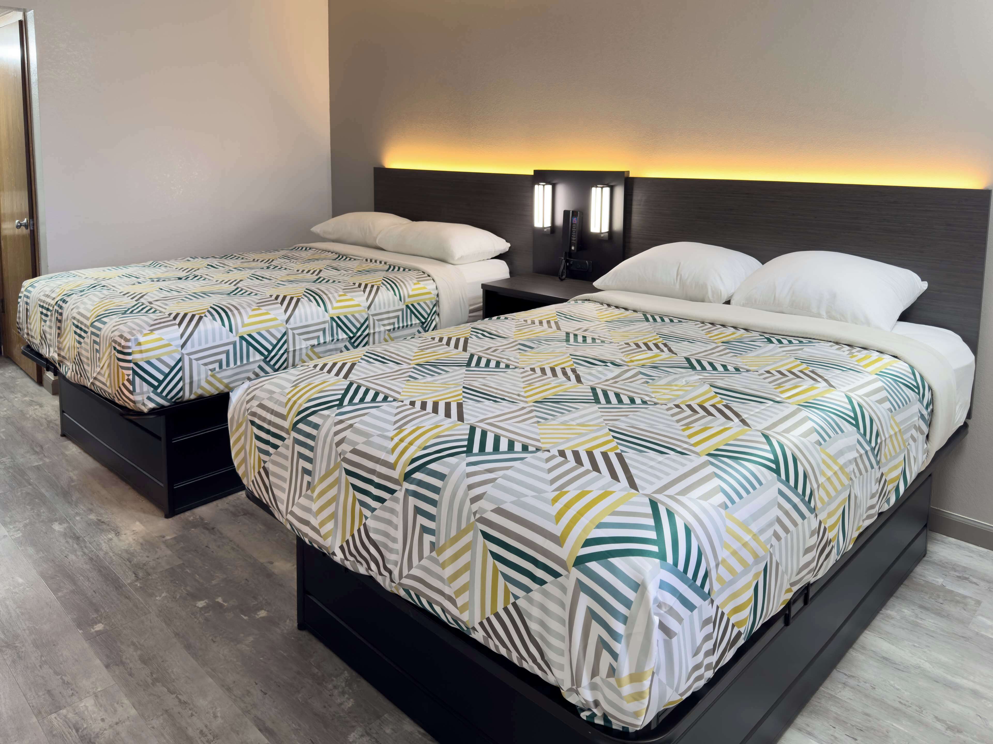A hotel room with two beds, each with a patterned bedspread and white pillows. There are wall-mounted lights above the beds and a wooden floor.
