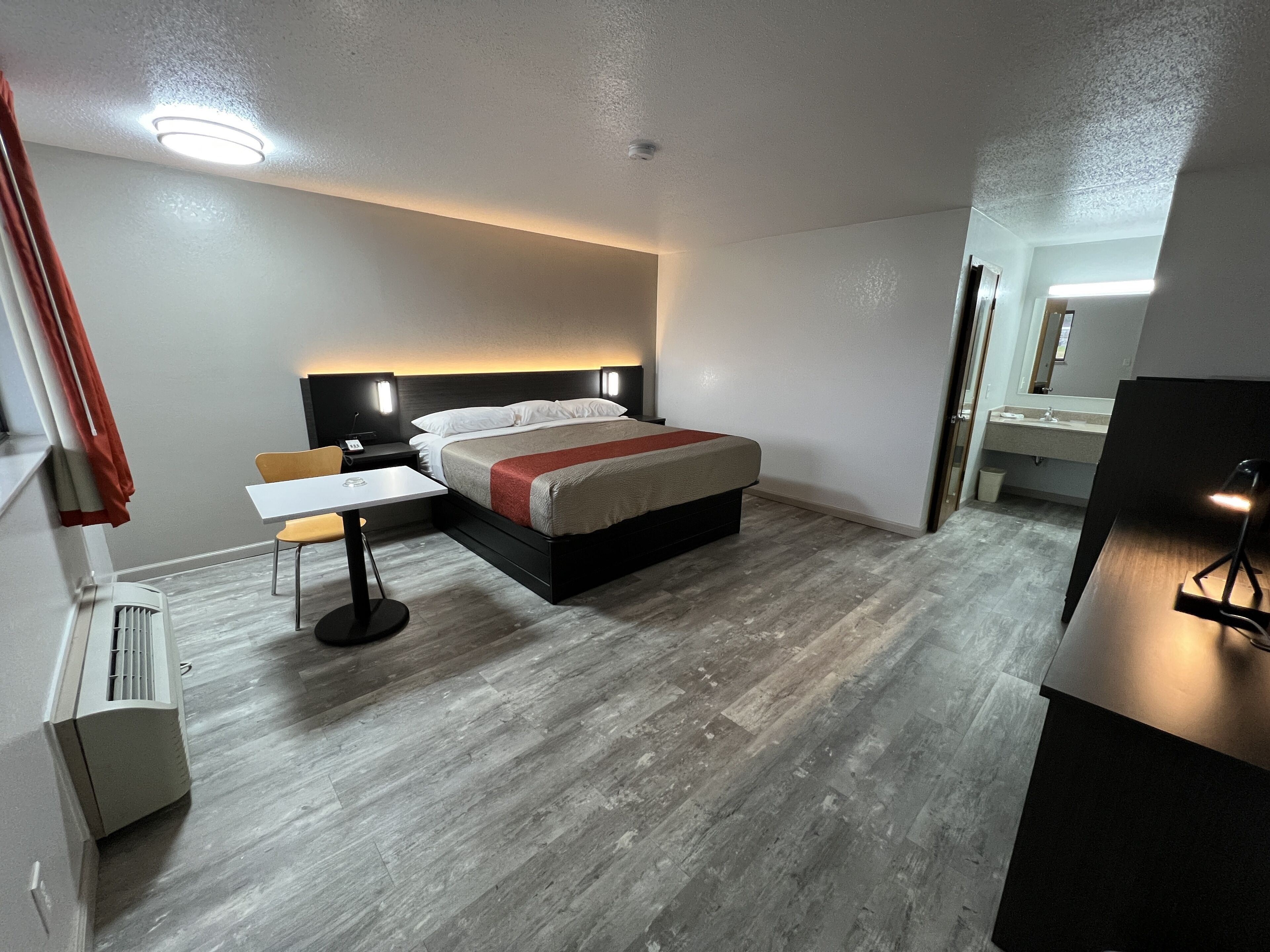 Una habitación de hotel moderna con cama, mesita de noche, silla y un pequeño escritorio con lámpara.