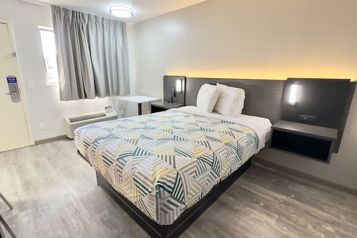 A modern hotel room with a large bed, a bedside table, a small desk, and a chair.