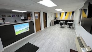 A hotel reception area with a desk, chairs, a small kitchenette, and a large screen displaying a message. - Motel 6 Dyersburg, TN (Dyersburg)