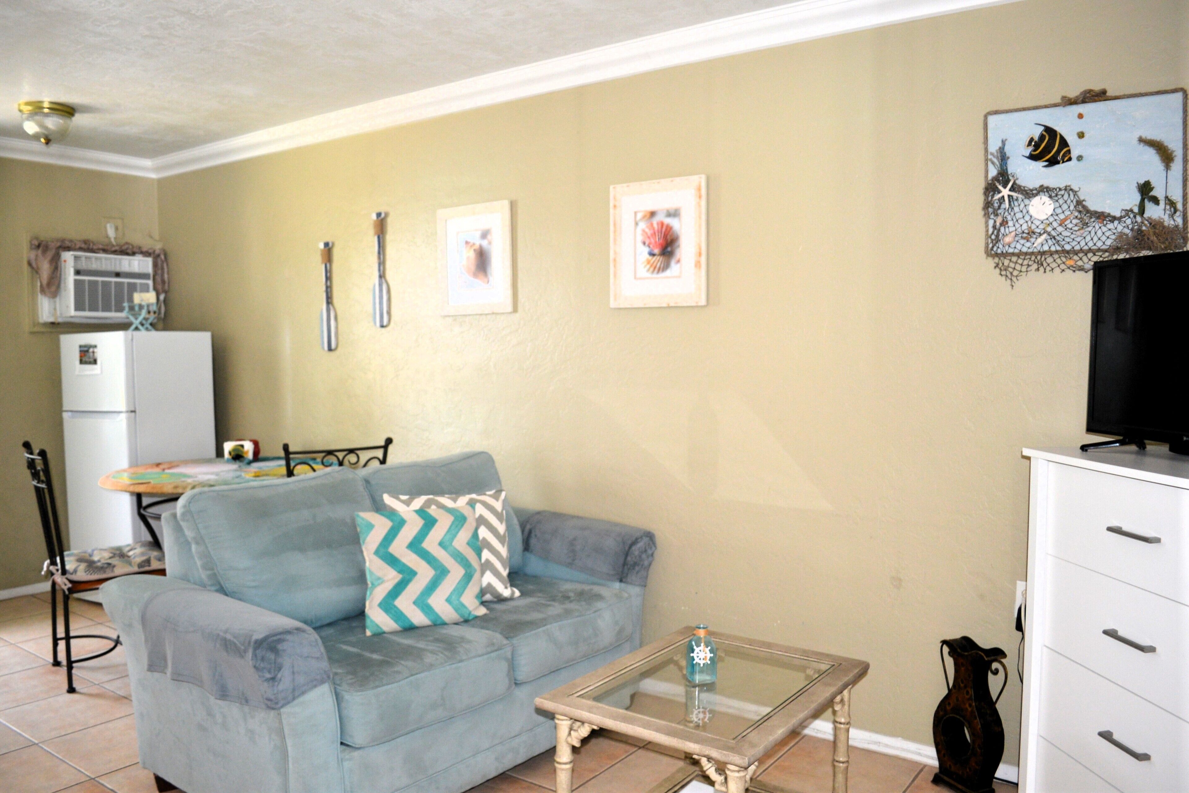 A living room with a sofa, a coffee table, a TV stand, and a kitchen area in the background.