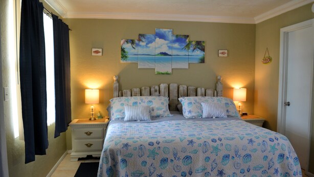 A bedroom with a bed, two bedside tables, a nightstand, a painting on the wall, and a door. - Sun Life Beach Hotel (Englewood)