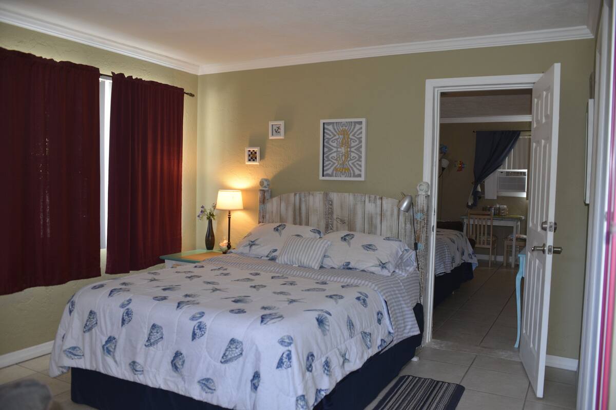 A bedroom with a large bed, a bedside table, a lamp, and a door leading to another room.