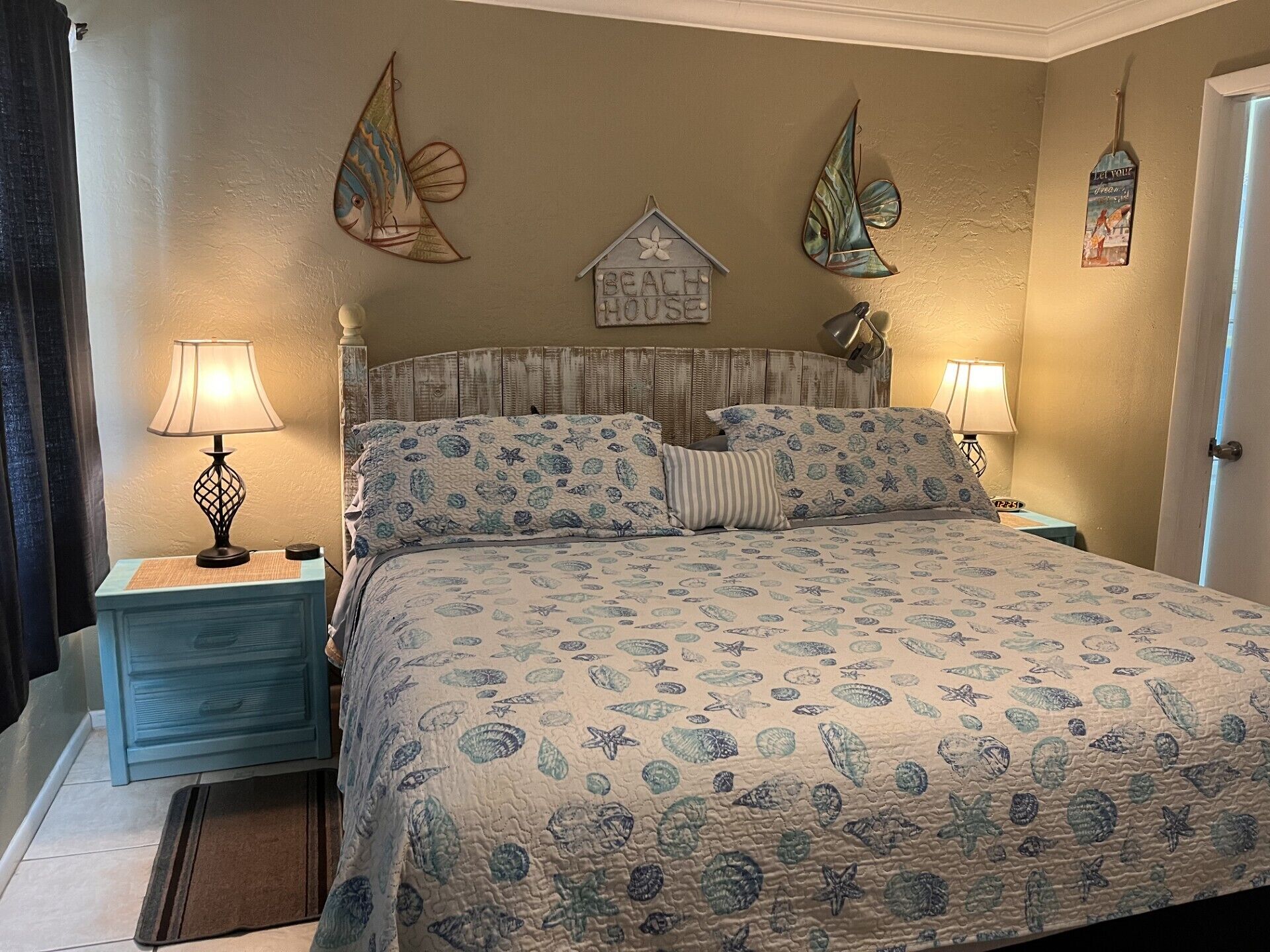 Un dormitorio con cama, dos mesitas de noche, una mesita auxiliar, un adorno en la pared y una puerta.