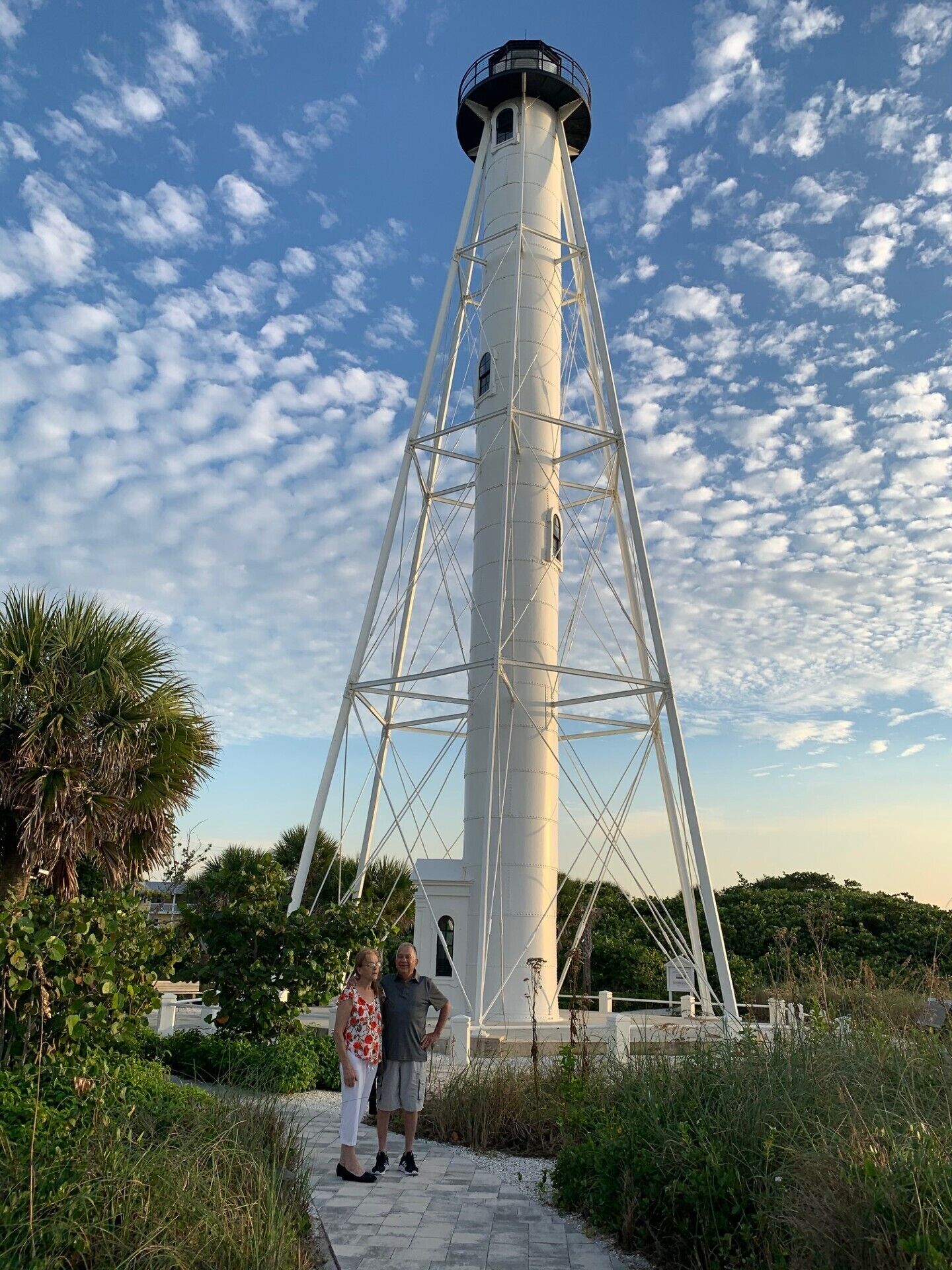 Un grand phare blanc, doté d’une structure métallique, entouré de verdure et surplombant un ciel dégagé.