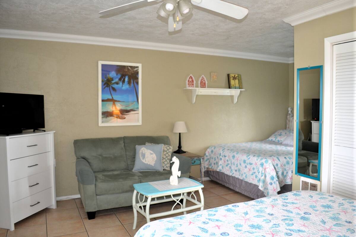 A bedroom with a bed, a sofa, a TV, a ceiling fan, and a painting of a beach scene.