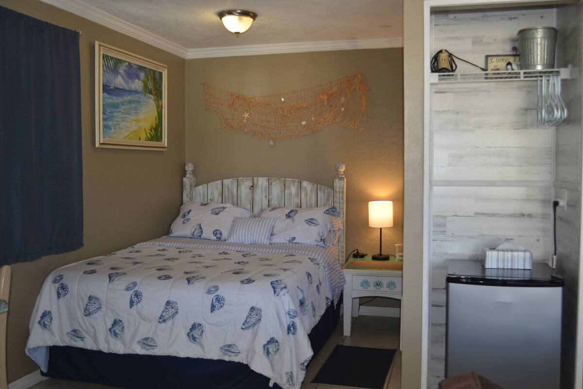A bedroom with a bed, a nightstand, a lamp, a refrigerator, and a painting on the wall.