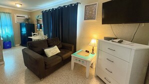 A compact living room with a sofa, a small table with a lamp, a TV mounted on the wall, and a white cabinet.