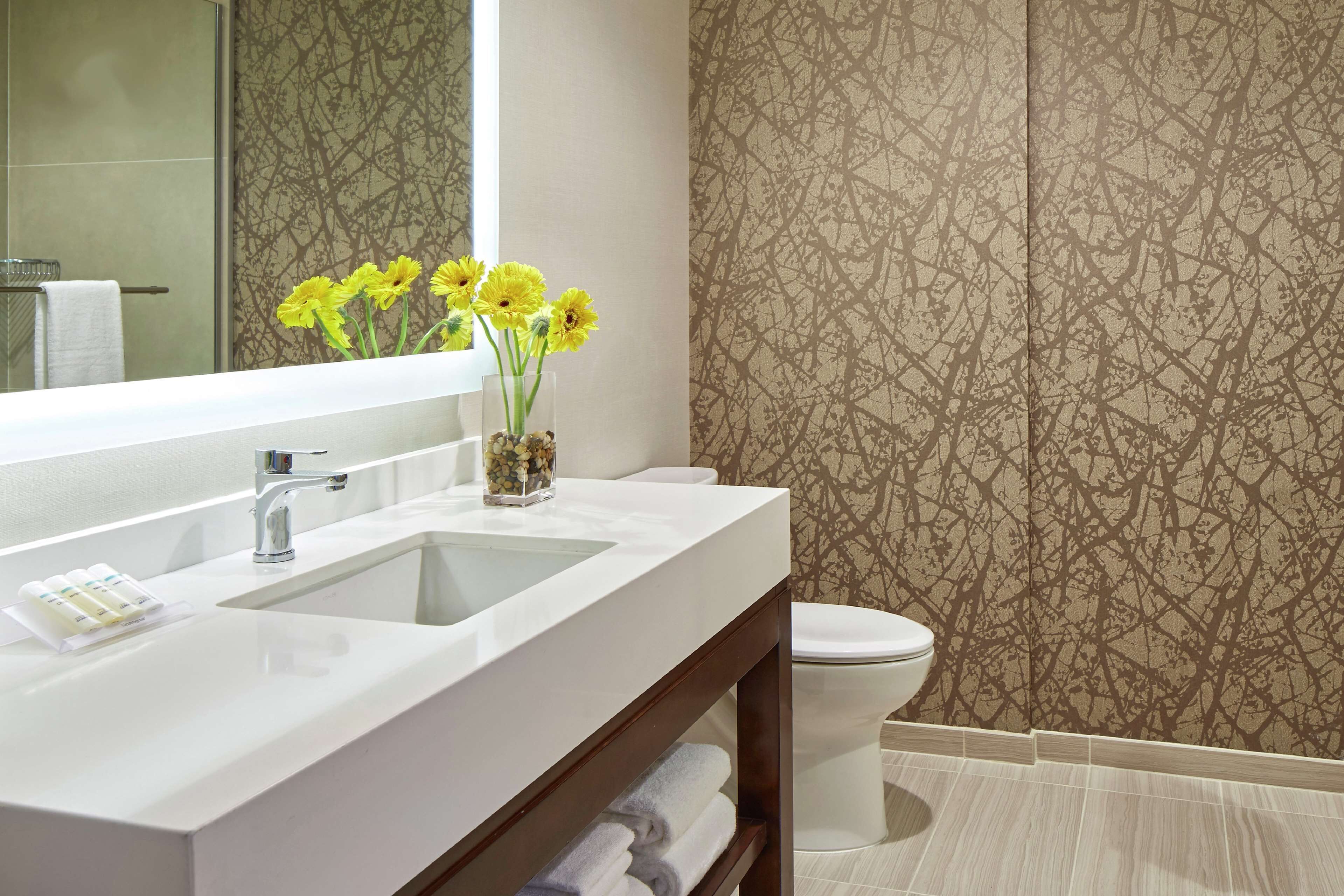A modern bathroom with a white sink, mirror, and a vase of yellow flowers. There is a toilet and a towel rack visible.