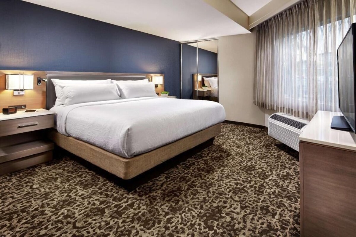 Une chambre d’hôtel comprenant un lit, des tables de chevet, un bureau, un miroir et une grande fenêtre avec des rideaux.