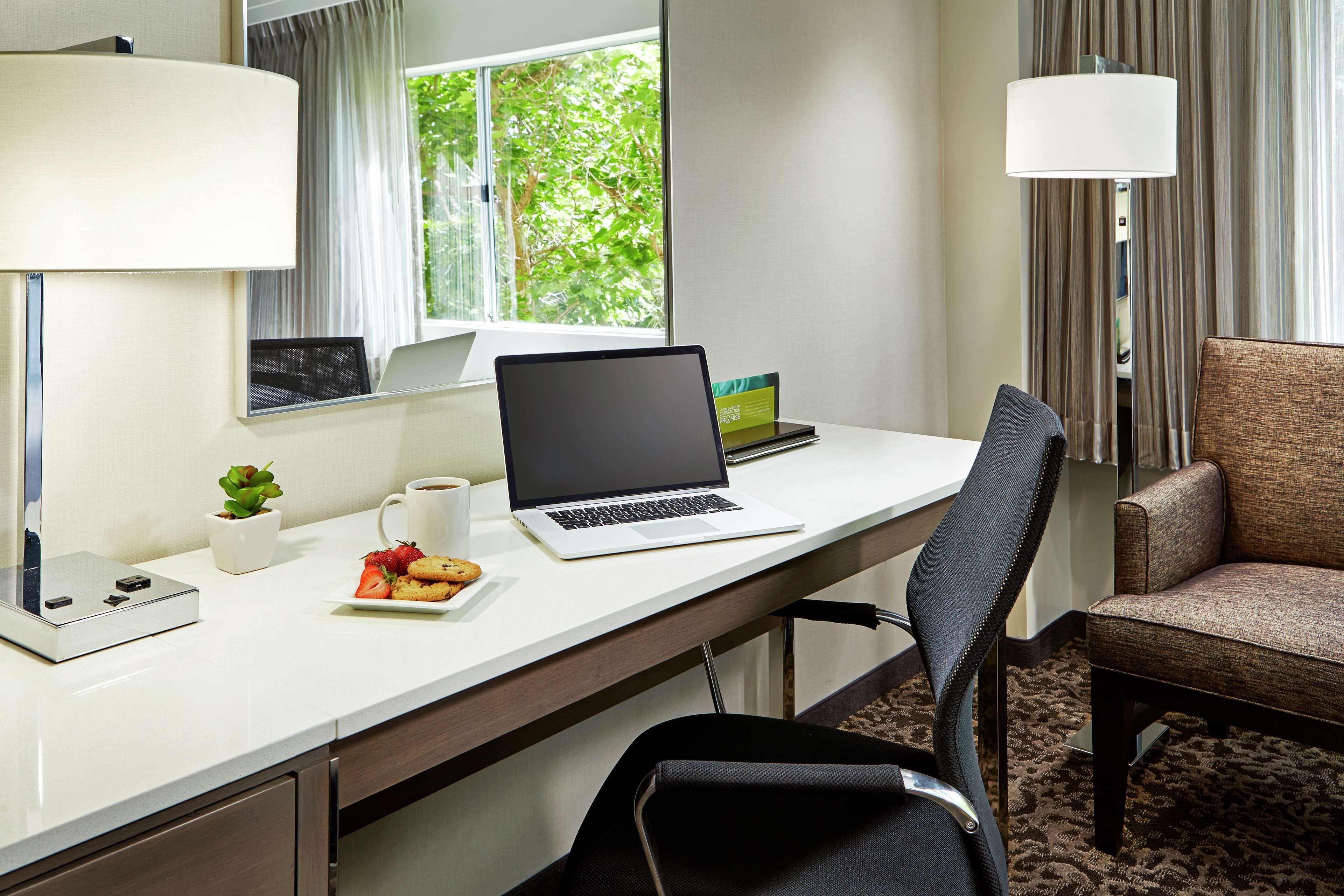 A hotel room with a desk, laptop, coffee cup, plate of cookies, and a chair.