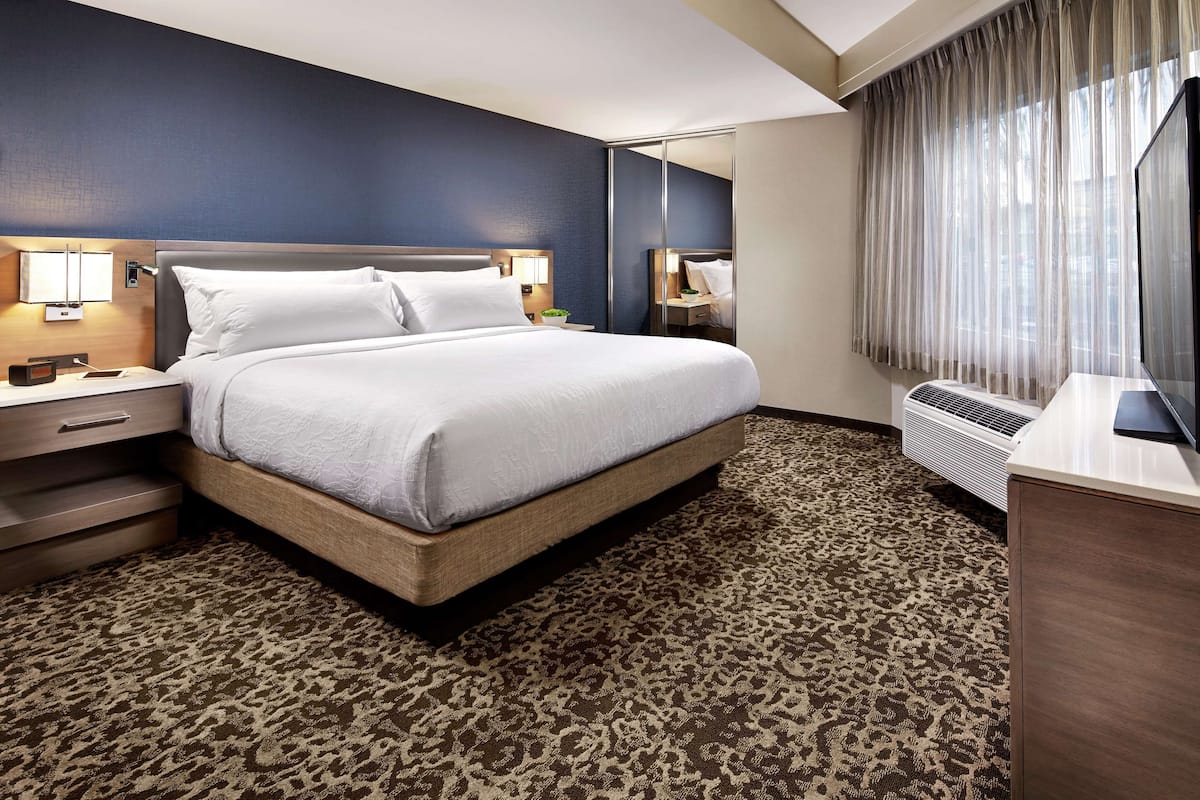 Ein Hotelzimmer mit Bett, Nachttischen, einem Schreibtisch und einem großen Fenster mit Vorhängen.