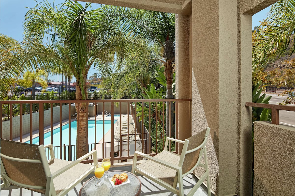 Ein Balkon mit zwei Stühlen, einem kleinen Tisch mit einem Getränk und einem Teller Essen, und Blick auf einen Swimmingpool und Palmen.