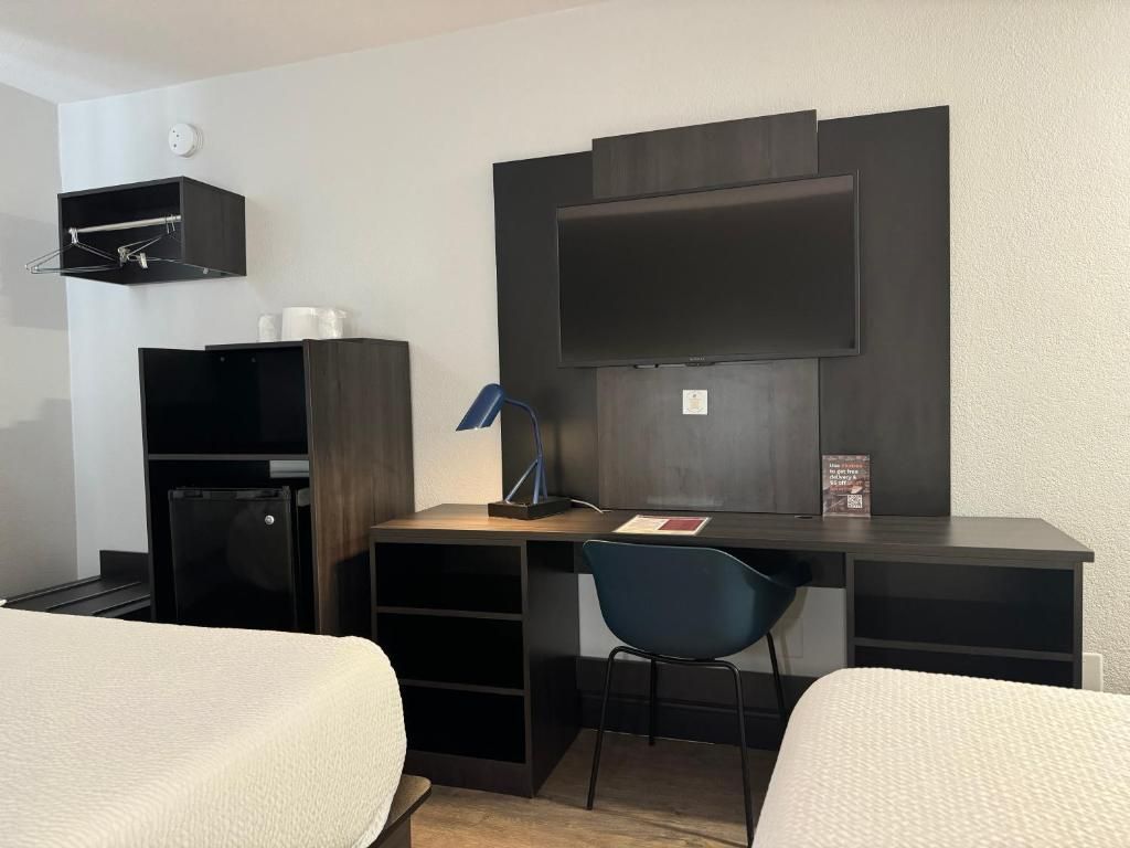 A hotel room with a bed, a desk with a chair, a TV mounted on the wall, and a small shelf with toiletries.