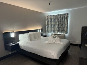 A neatly made bed with white linens and pillows, a bedside table with a lamp, and a patterned curtain on the window.