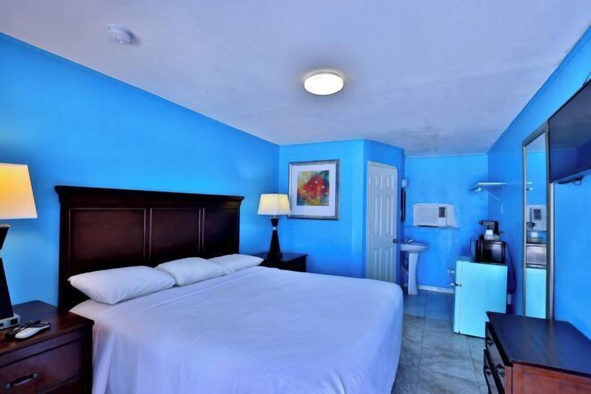 A hotel room with a large bed, bedside tables, a nightstand, a desk, a chair, a mirror, and a door.