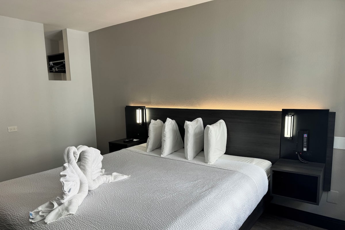 A neatly made bed with white linens and pillows, a bedside table with a phone and a lamp, and a wall-mounted shelf above the bed.