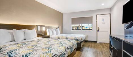 A hotel room with two beds, a desk, a TV, and a window with a view of buildings.