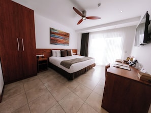 A hotel room with a bed, a desk, a wardrobe, a ceiling fan, a painting, and a window with curtains. - Hotel Cucuve (Puerto Ayora)