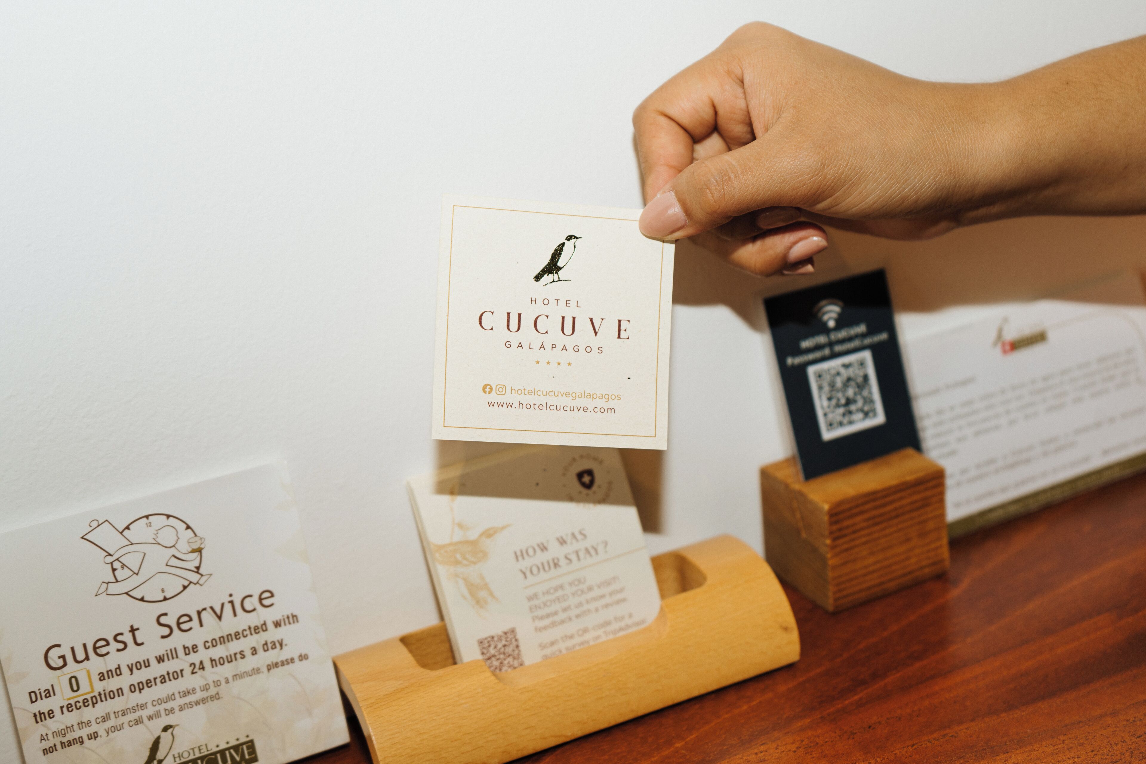Una mano sostiene una tarjeta de presentación del Hotel Cucuve, Galápagos, con un código QR y una tarjeta de servicios para huéspedes debajo.