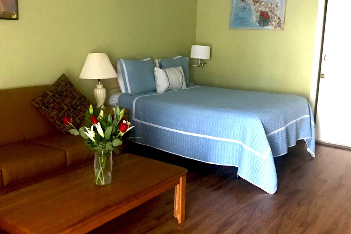 A room with a bed, a wooden bench, a table with a vase of flowers, a ceiling fan, and a painting on the wall.