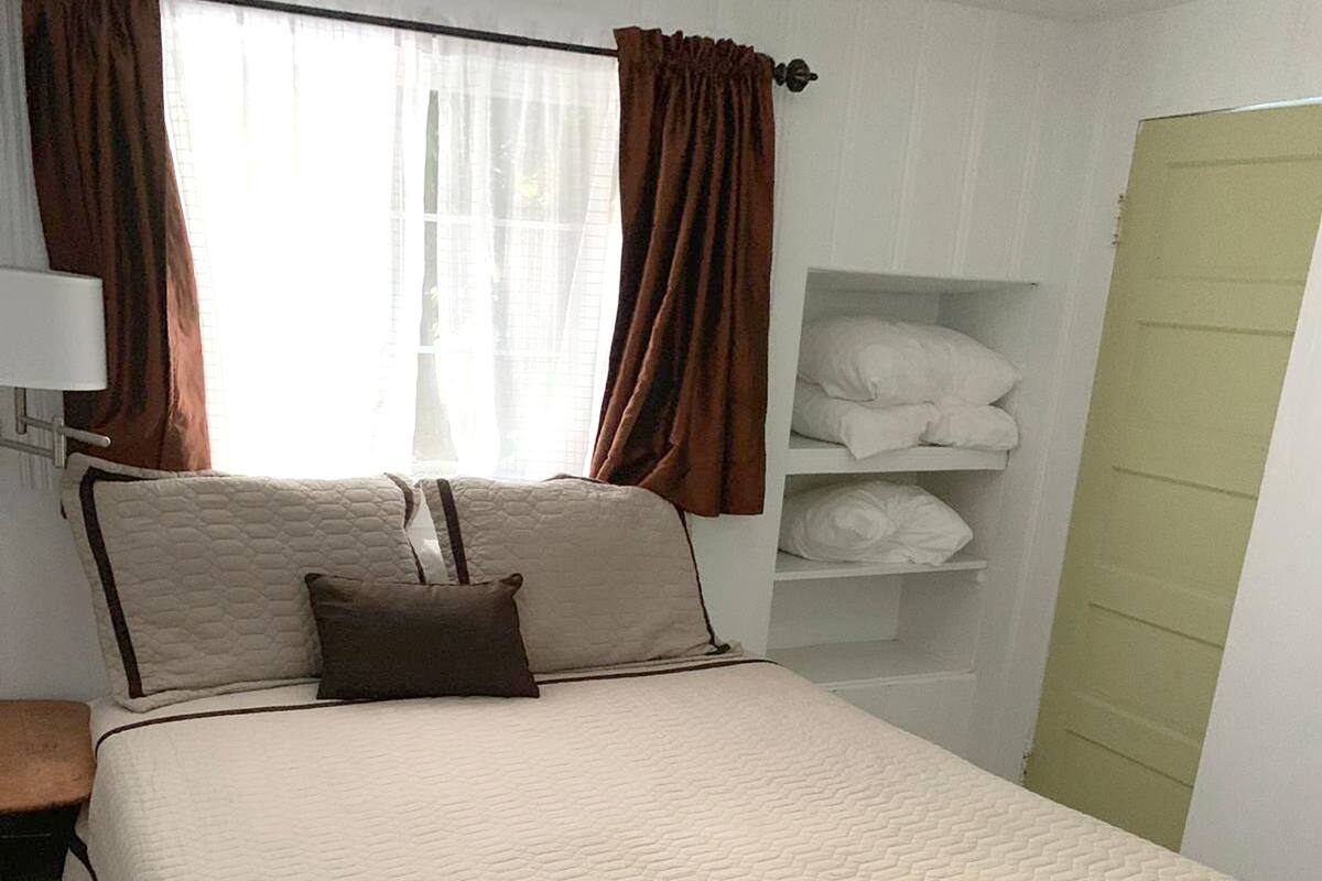 A neatly made bed with a quilted cover, a bedside table, a shelf with folded linens, and a window with curtains.