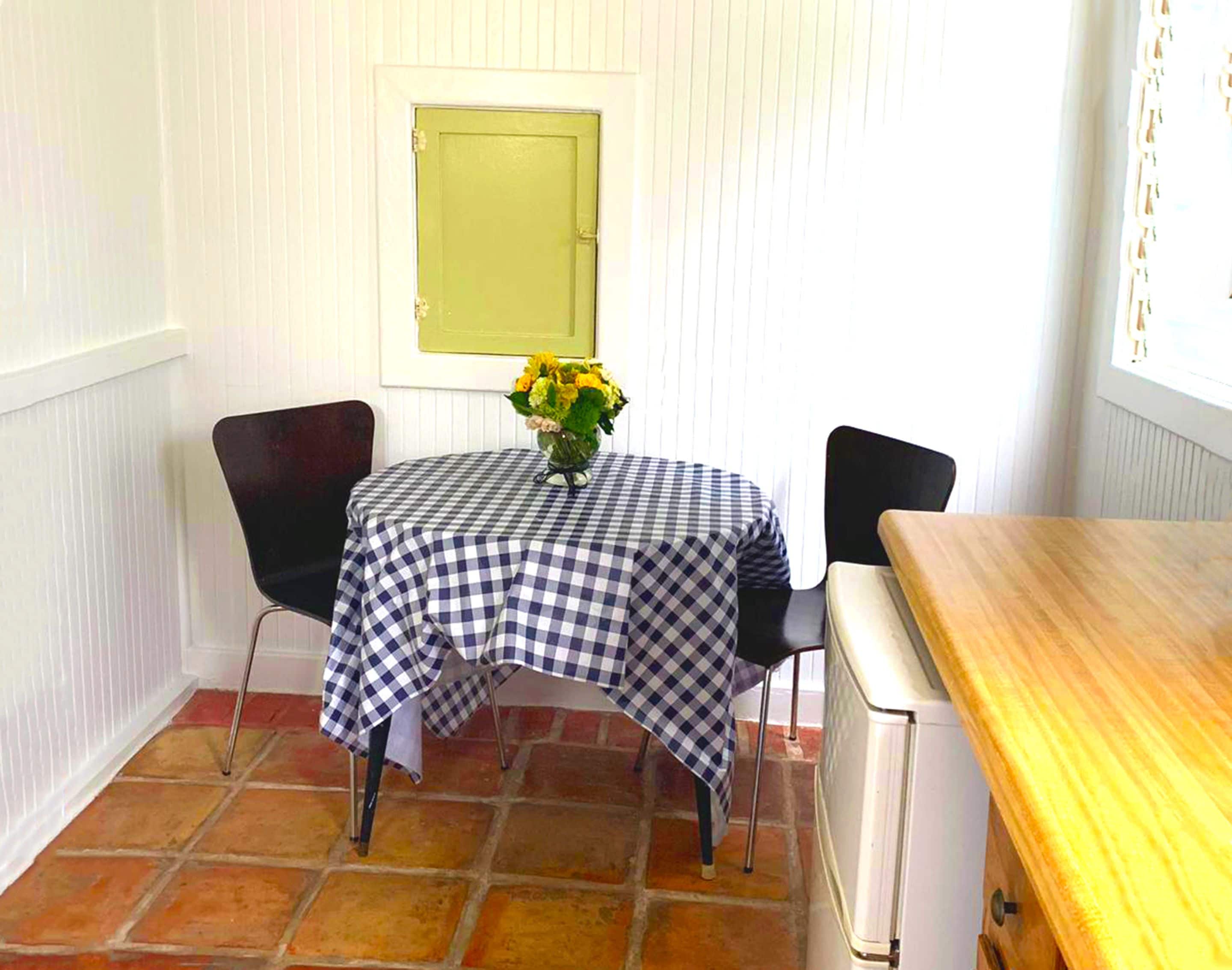 Un pequeño comedor con mantel a cuadros, dos sillas, un jarrón con flores y una encimera de madera.