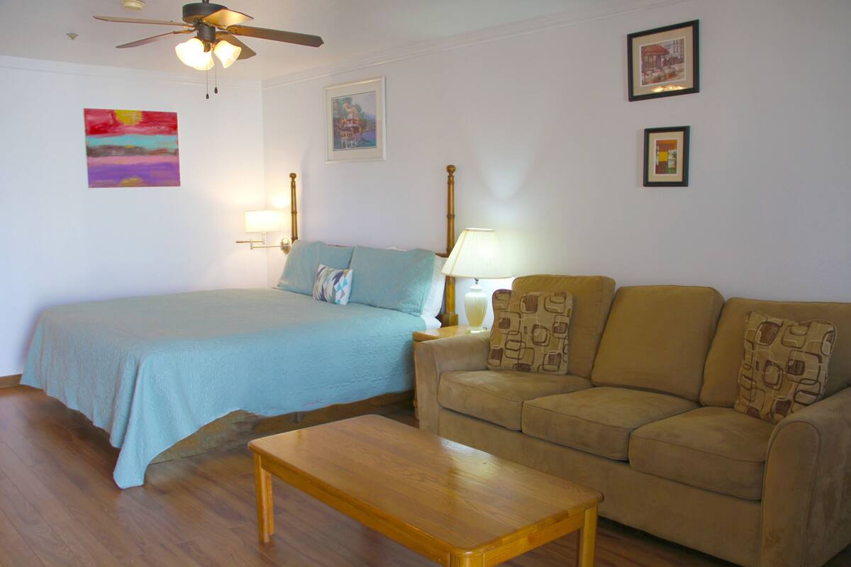A bedroom with a bed, a wooden nightstand with a lamp, a beige sofa, and a wooden coffee table.