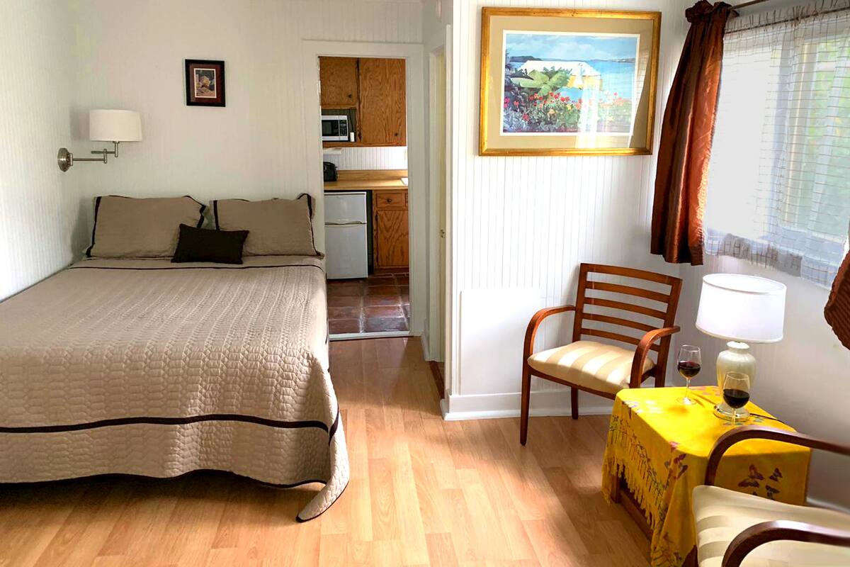 A bedroom with a bed, a chair, a table with drinks, a painting, and a kitchen area.