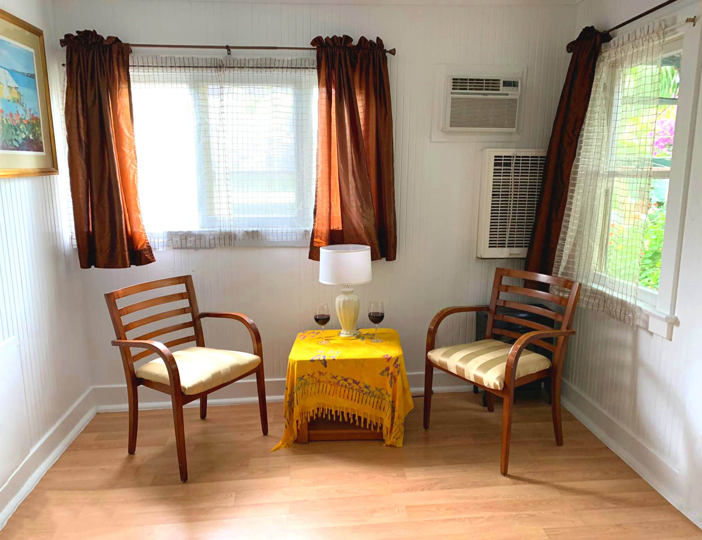 A room with two wooden chairs, a small table with a yellow tablecloth, and a lamp. There are windows with curtains, an air conditioning unit, and a painting on the wall.