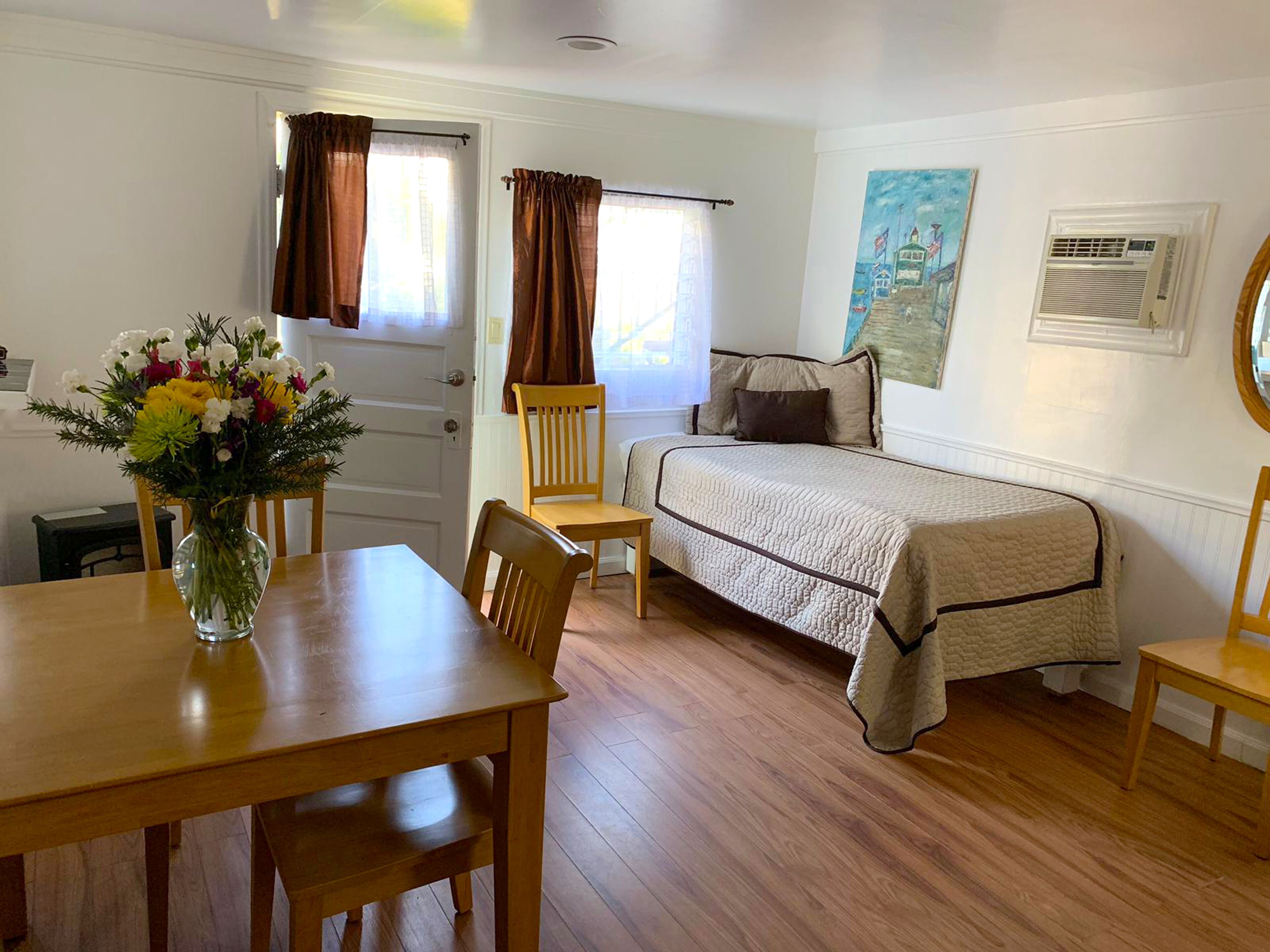 A room with a wooden table, a bed with a quilt, a chair, a vase with flowers, and a painting on the wall.
