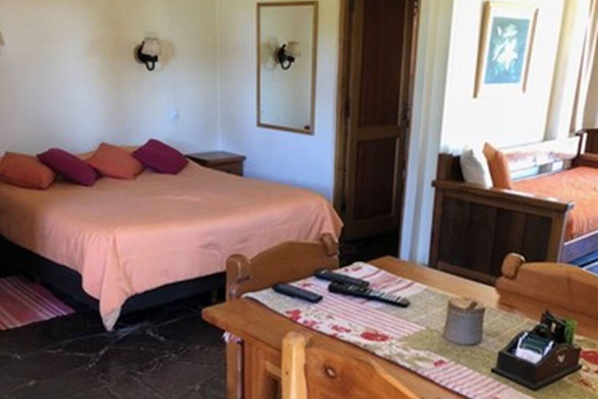 A room with a bed, a wooden table with chairs, a small table with a lamp, and a door.
