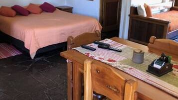 A room with a bed, a wooden table with chairs, a small table with a lamp, and a door.