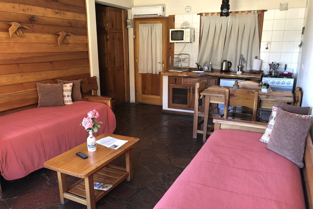 A cozy room with two beds, a wooden table with a vase of flowers, a small kitchen area with a microwave and stove, and a wooden wardrobe.