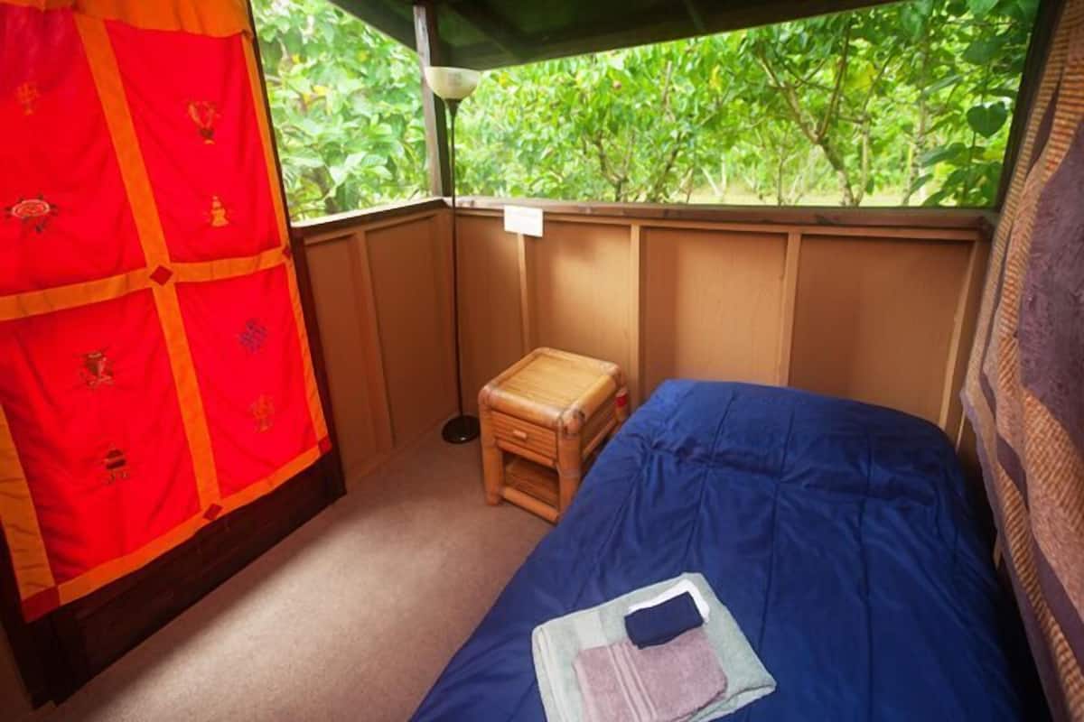 Une petite pièce fermée, avec un lit simple, un petit tabouret en bois et une fenêtre donnant sur la verdure.