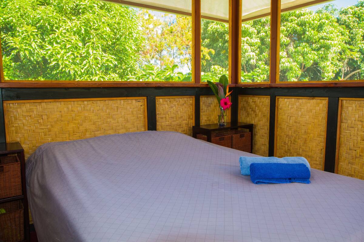 Une pièce avec un lit, une fenêtre donnant sur un paysage verdoyant, un mur recouvert d’un revêtement tissé et une petite table sur laquelle est posé un vase.