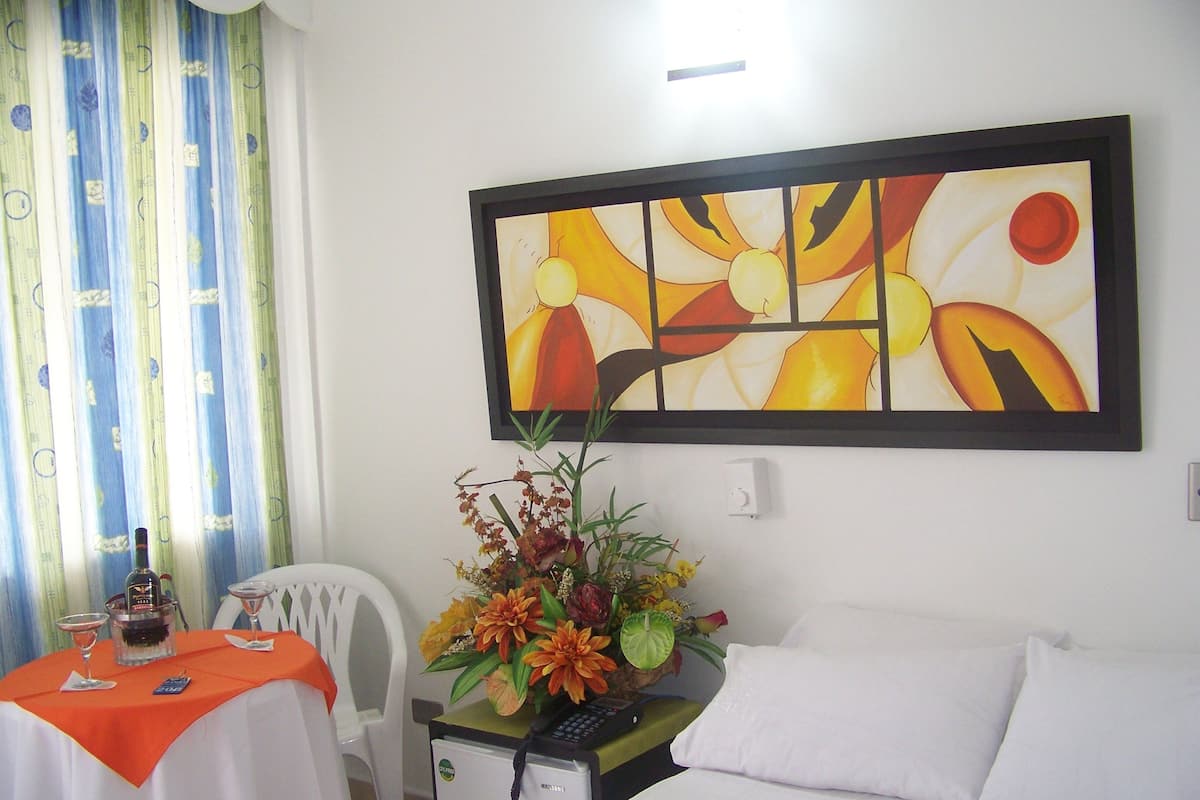 A hotel room with a bed, a desk with a computer, a chair, a table with a bottle and glasses, and a painting on the wall.