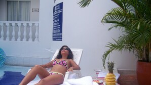 A person relaxing on a sun lounger by a pool, with a sign indicating pool rules and a potted plant nearby.