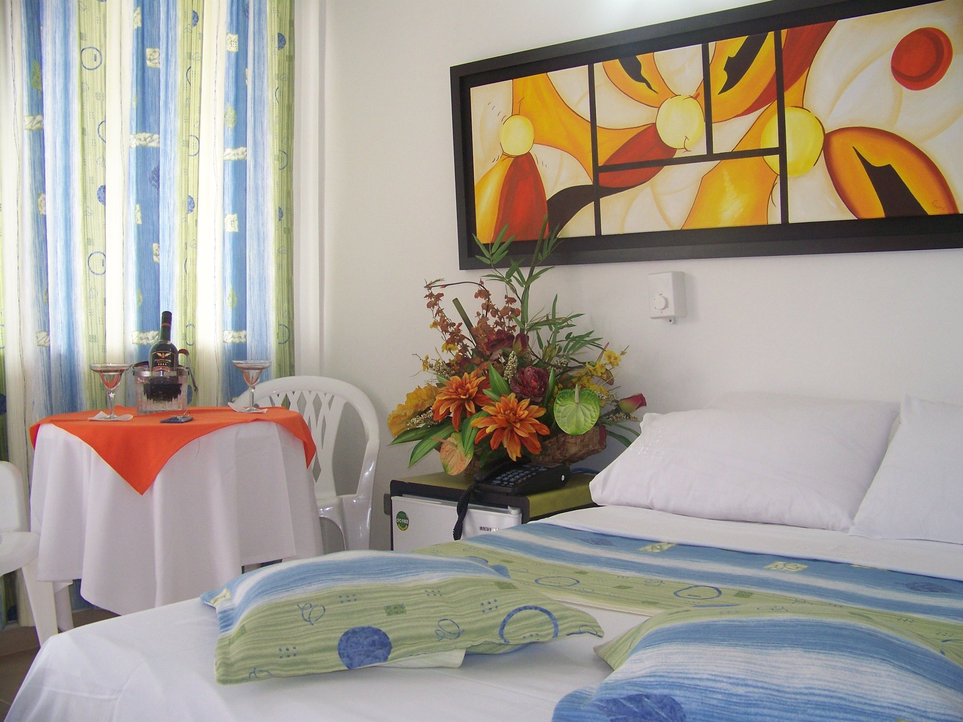 Quarto de hotel com uma cama, uma mesa com garrafa e copo, uma cadeira, um quadro e um vaso com flores.