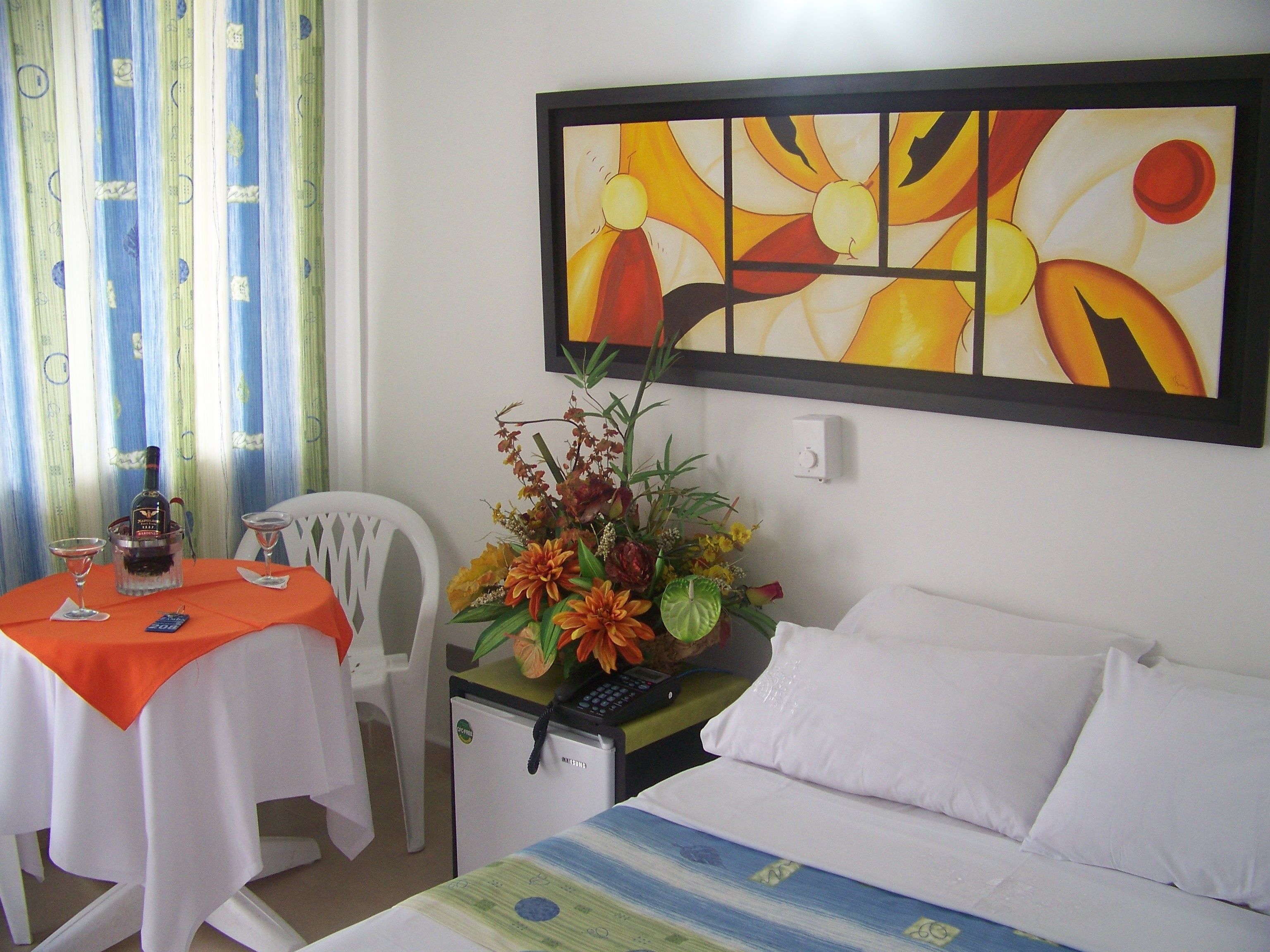 A hotel room with a bed, a small desk with a chair, a table with a bottle and glasses, a refrigerator, and a painting on the wall.