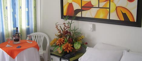 A hotel room with a bed, a small desk with a chair, a table with a bottle and glasses, a refrigerator, and a painting on the wall.